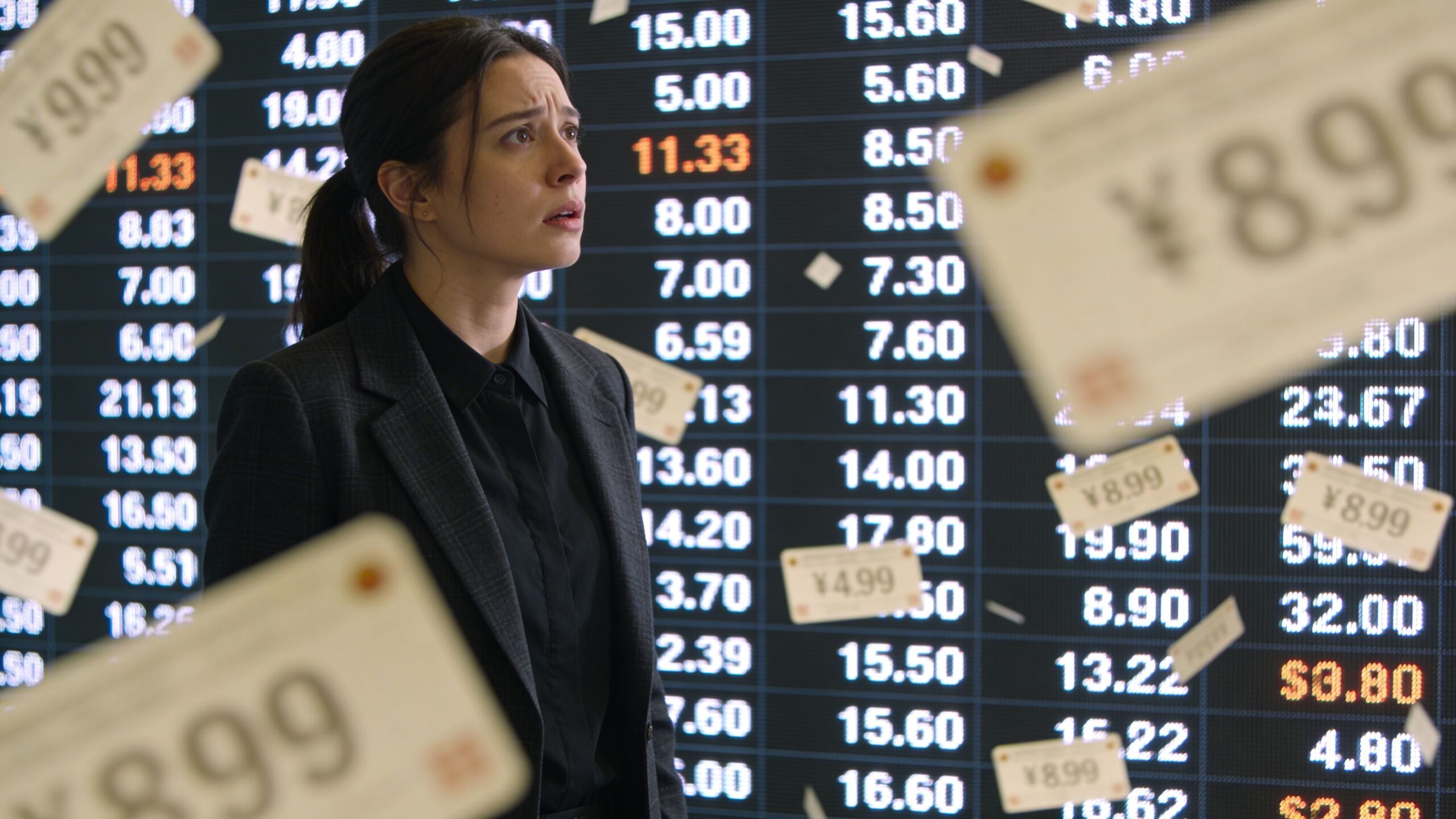 A concerned professional woman standing in front of a digital LED video wall displaying fluctuating market prices.